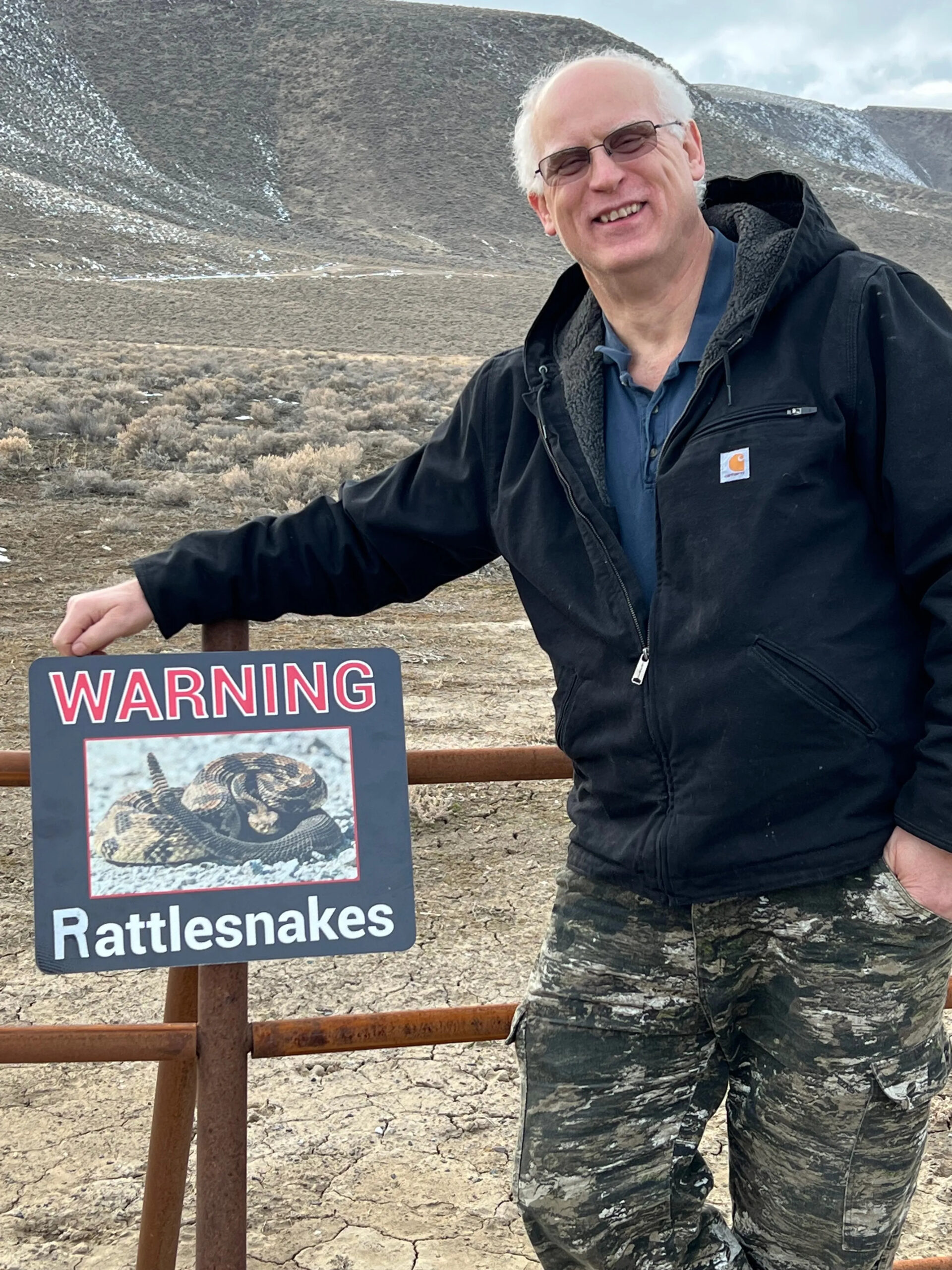 Man in a black jacket leans against a fence beside a "Warning: Rattlesnakes" sign, set in a dry, hilly landscape.