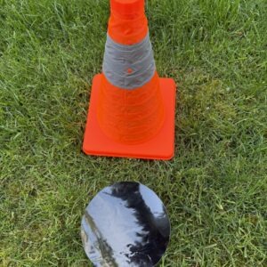 An orange traffic cone stands on green grass beside a shiny black circular object. The setup suggests a DIY project or safety demonstration.
