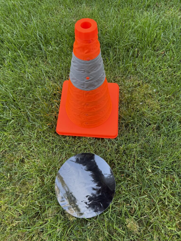 An orange traffic cone stands on green grass beside a shiny black circular object. The setup suggests a DIY project or safety demonstration.
