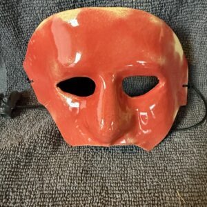 Red ceramic mask with a pronounced nose and two eye holes, showcasing a glossy finish. Displayed against a textured gray background.