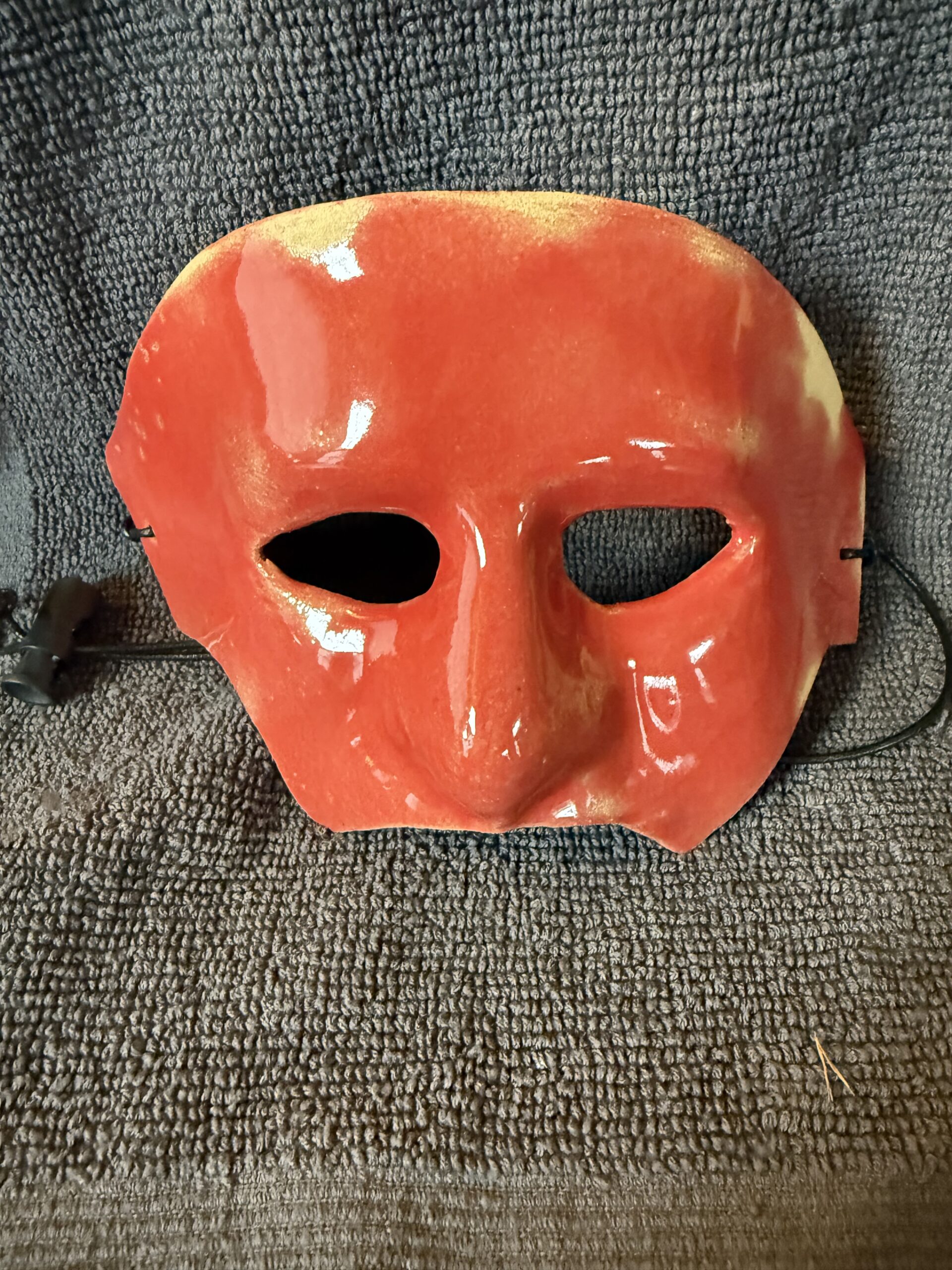 Red ceramic mask with a pronounced nose and two eye holes, showcasing a glossy finish. Displayed against a textured gray background.