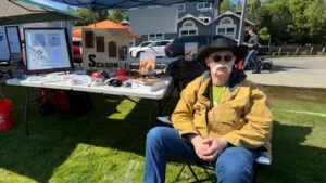 A man in a cowboy hat sits at a craft fair booth, showcasing books and handmade items, with a sunny park setting in the background.