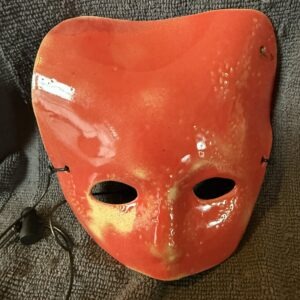 Ceramic mask in vibrant red with two eye holes, displayed against a textured gray background, highlighting its artistic craftsmanship.