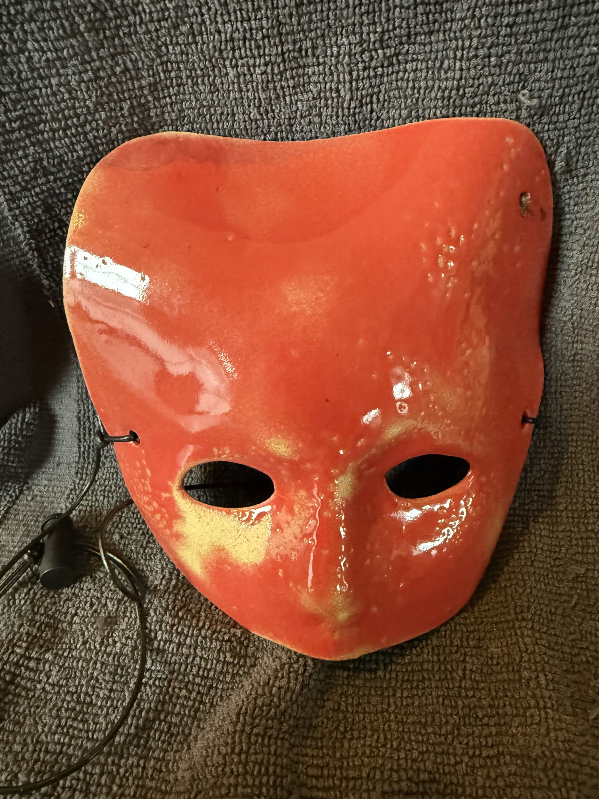 Ceramic mask in vibrant red with two eye holes, displayed against a textured gray background, highlighting its artistic craftsmanship.
