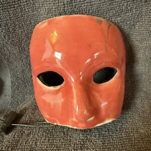 Ceramic mask in vibrant red with two eye holes, displayed against a textured gray background, highlighting its artistic craftsmanship.