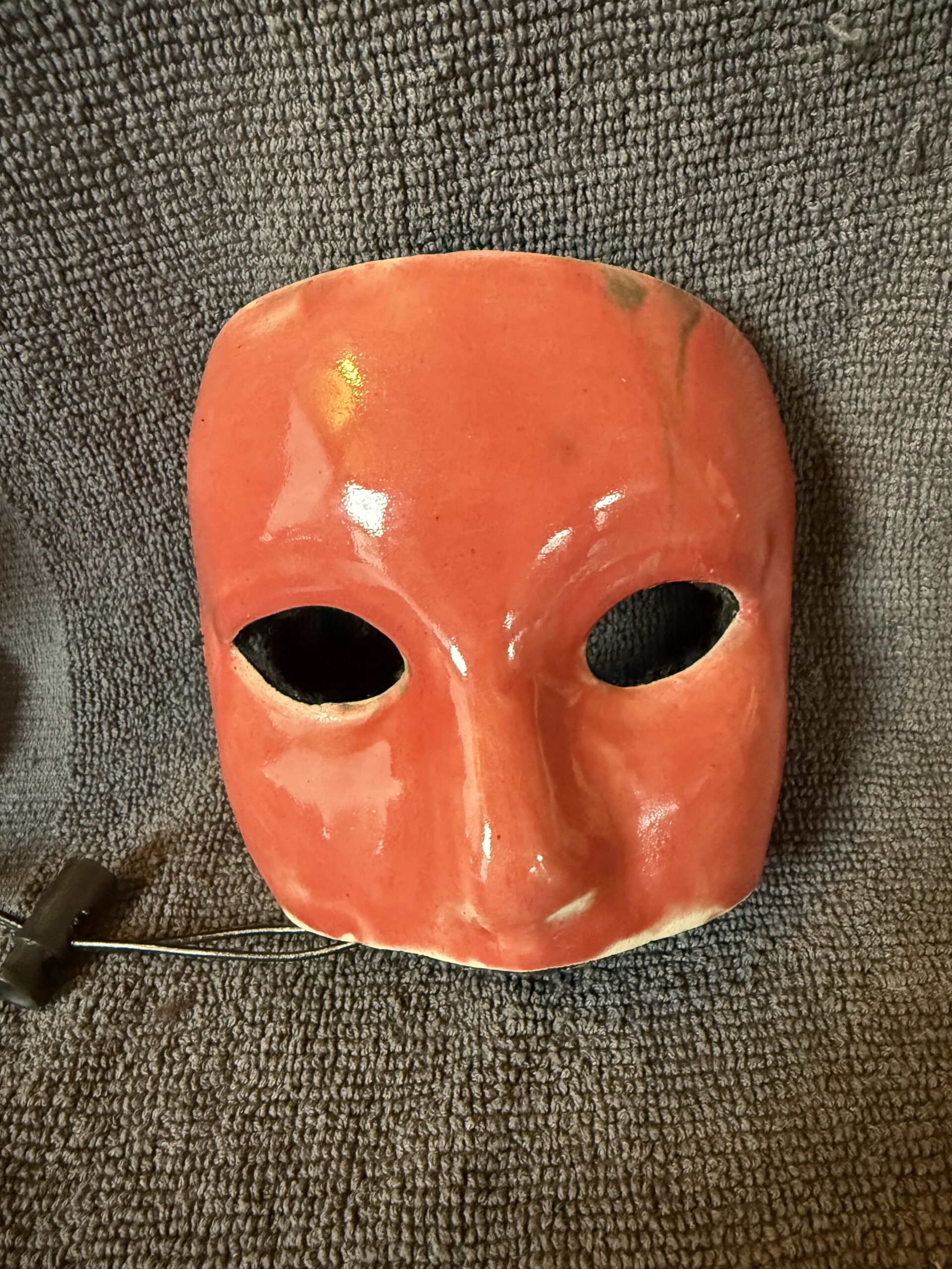 Ceramic mask in vibrant red with two eye holes, displayed against a textured gray background, highlighting its artistic craftsmanship.