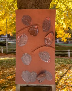 Ceramic artwork on a tree trunk featuring textured clay leaves in various colors against a backdrop of vibrant fall foliage.