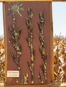 Artwork titled "The Seasons" depicts stylized corn stalks with a sun, representing agricultural themes. Installed outdoors near a cornfield.