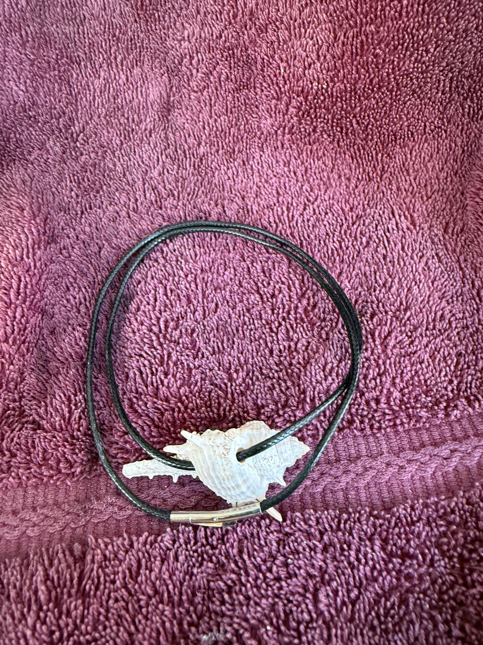 Black cord necklace featuring a white shell pendant, resting on a textured purple towel. Ideal for showcasing beach-themed fashion accessories.