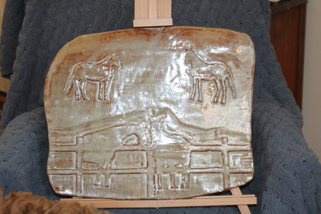 Cream-glazed handmade clay wall hanging showing horses standing in a fenced pasture